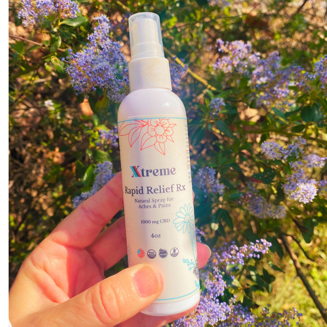 Xtreme Brands RX Rapid Relief Spray held in front of garden flowers – strongest doctor-trusted formula with full-spectrum hemp, Arnica, and Menthol for fast, hands-free pain relief.