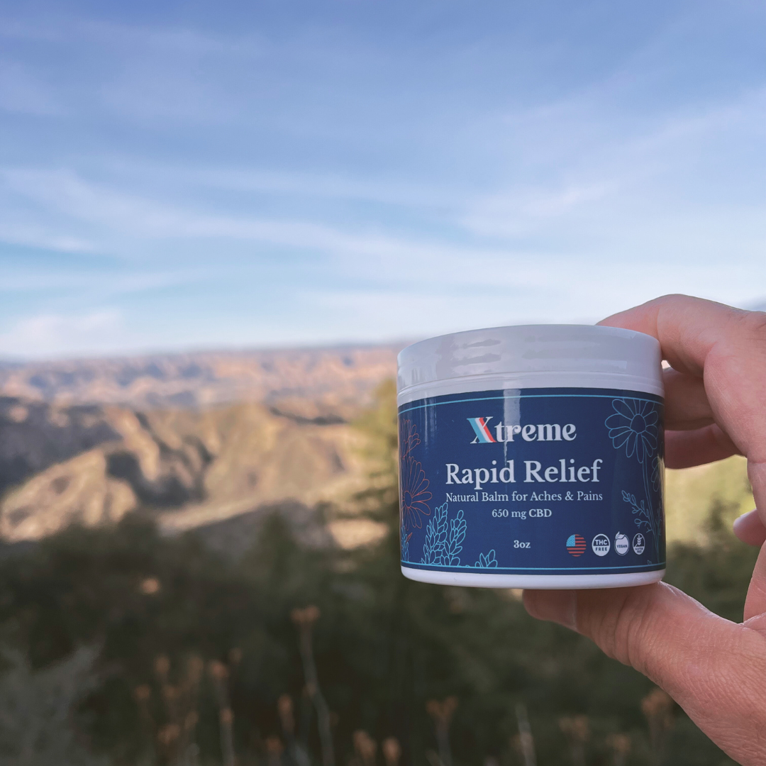 Xtreme Rapid Relief Natural Balm held up in a nature setting – USA-made, doctor-trusted hemp-derived CBD formula with Arnica, Turmeric, and Willow Bark for everyday muscle and joint relief.