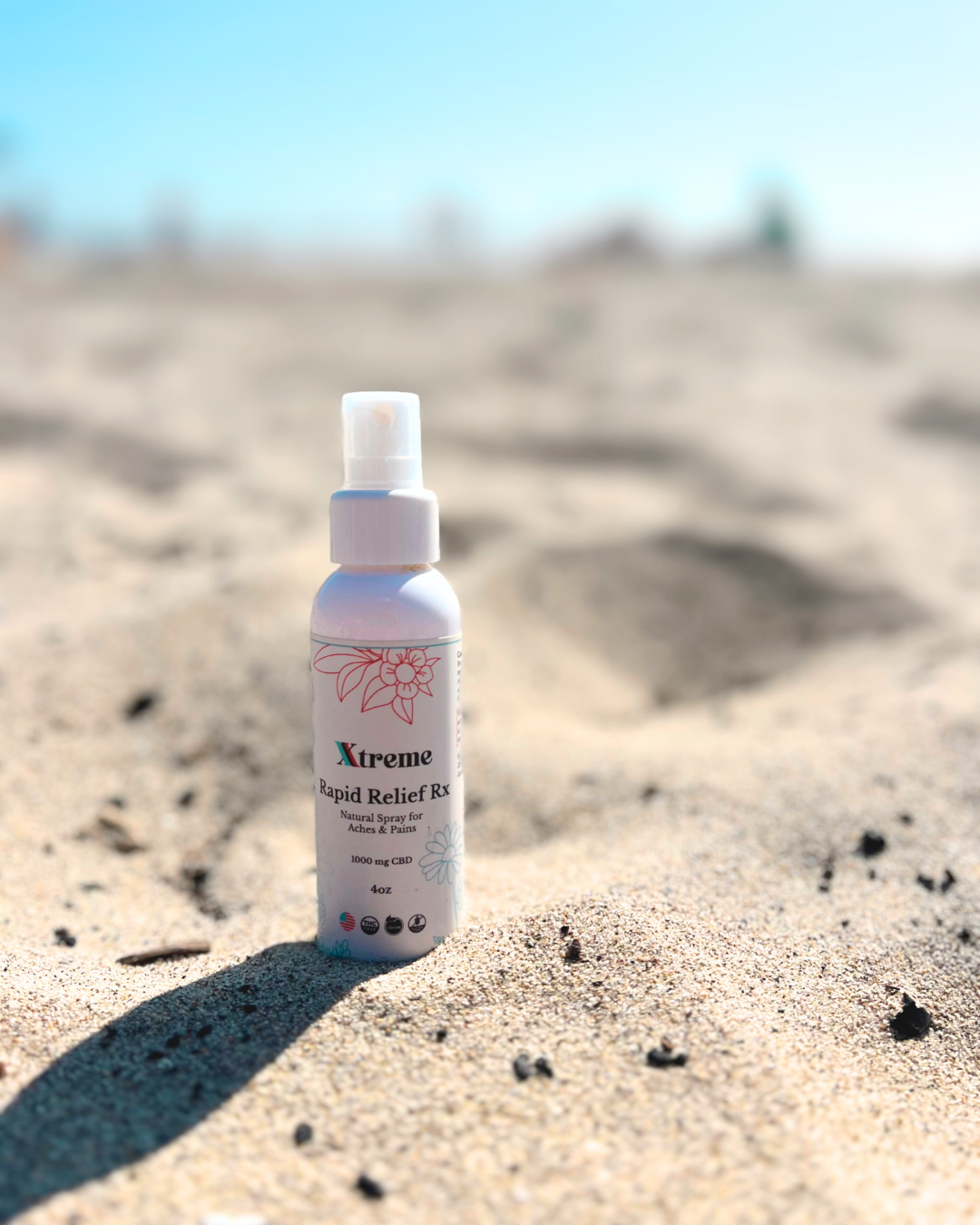 Xtreme Brands RX Rapid Relief Spray sitting in the sand – strongest doctor-trusted formula with full-spectrum hemp, Arnica, and Menthol for fast, hands-free pain relief.
